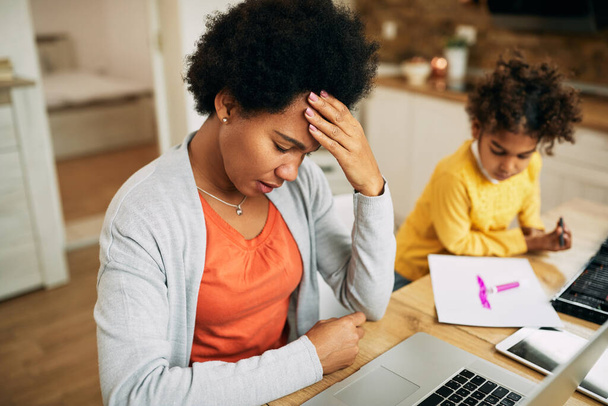 Афроамериканская мать чувствует стресс и держит голову в боли во время работы за компьютером дома.  - Фото, изображение