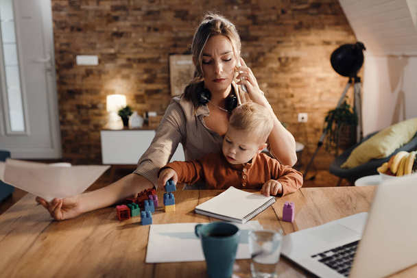 Single mother communicating over mobile phone while babysitting her son and working at home.  - Foto, Bild
