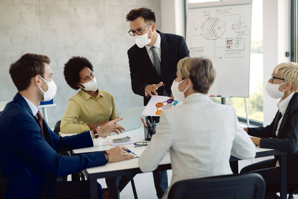 Young entrepreneur and his colleagues brainstorming while working on new business strategy and examining charts on a meeting. They are wearing protective face masks due to coronavirus pandemic.  - 写真・画像
