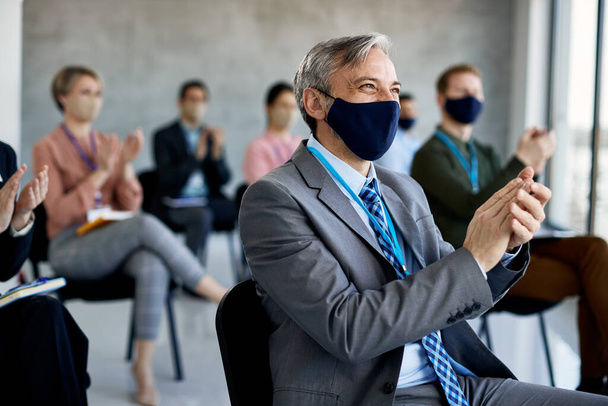 Ευτυχής επιχειρηματίας και οι συνάδελφοί του χειροκροτούν μετά από επιτυχημένο επιχειρηματικό σεμινάριο και φορώντας μάσκες προσώπου λόγω πανδημίας COVID-19. - Φωτογραφία, εικόνα