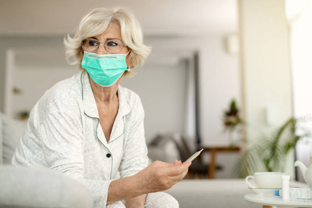 Mature woman using thermometer and measuring her temperature while wearing protective face mask at home during coronavirus pandemic.  - Photo, Image