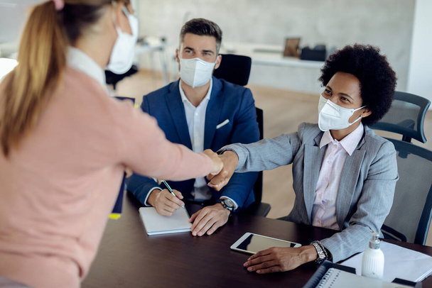 Members of human resource team wearing protective face masks while having job interview with potential candidate in the office.  Focus is on black businesswoman fist bumping with a candidate.  - Photo, image