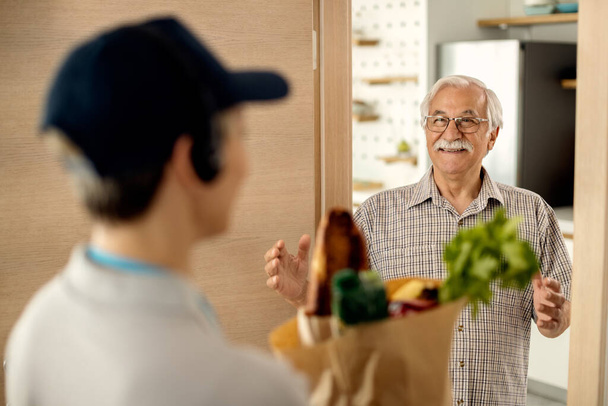 Щасливий старший чоловік розмовляє з визволителем, який приносить йому сумку з продуктами вдома
.  - Фото, зображення