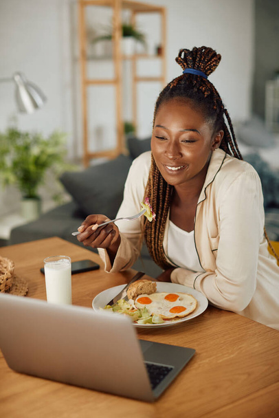 Happy black woman eating breakfast while making video call over laptop at home. - Foto, imagen