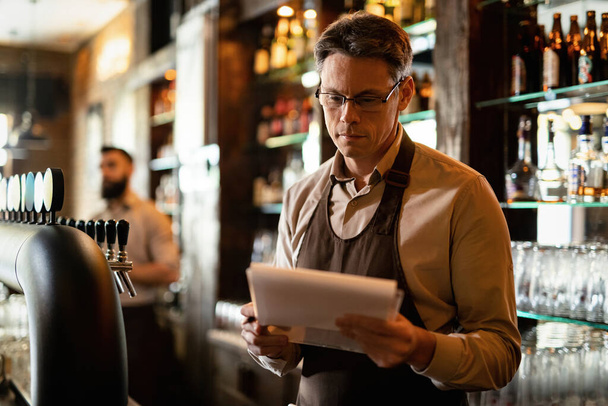 Усміхнений середній дорослий бармен проходить через список під час роботи в пабі
.  - Фото, зображення