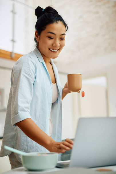 Genç Asyalı kadın sabah evde dizüstü bilgisayarla internette sörf yaparken çay içiyor..  - Fotoğraf, Görsel