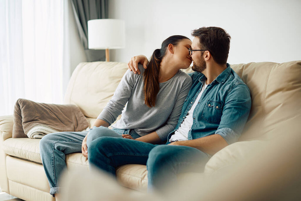 Закохана пара обіймає і цілує сидячи на дивані у вітальні
.  - Фото, зображення