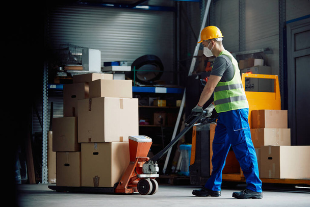 Warehouse worker with face mask pushing packages on pallet jack at storage compartment. - 写真・画像