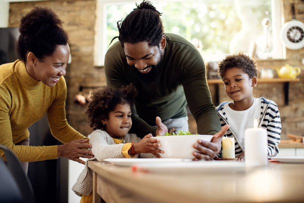 Happy black parents and their children bringing food at the table in dining room.  - Фото, изображение
