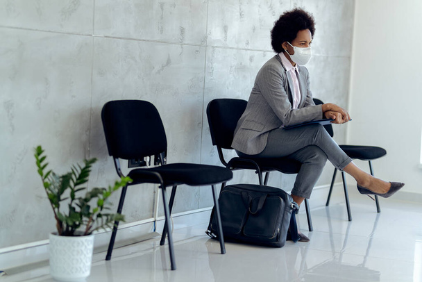 Distraught African American businesswoman wearing protective face mask while sitting in a hallway and waiting for job interview.  - Valokuva, kuva