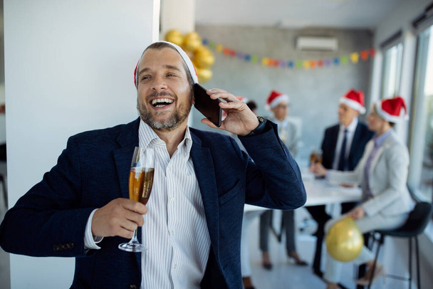 Mid adult businessman having fun while drinking Champagne and communicating on mobile phone during Christmas party in the office. His colleagues are in the background.  - Photo, Image