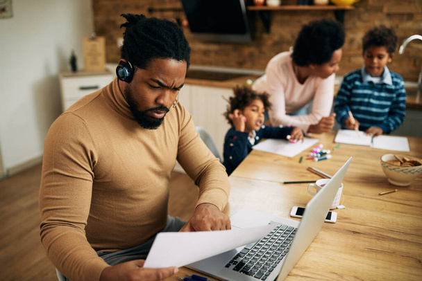 African American father working on a computer and going through reports while his wife is homeschooling their kids.  - Photo, image