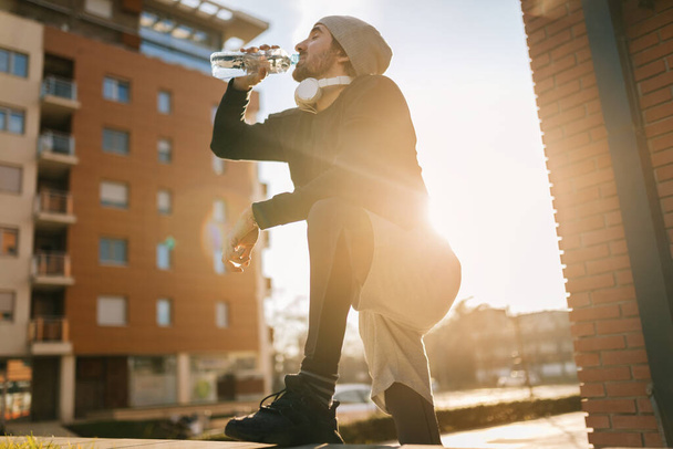 Low angle view of thirsty athletic man drinking water form a bottle while taking a break in the city.  - 写真・画像