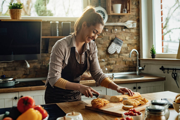 Young happy woman slicing baguette while preparing food in the kitchen. - Foto, Bild