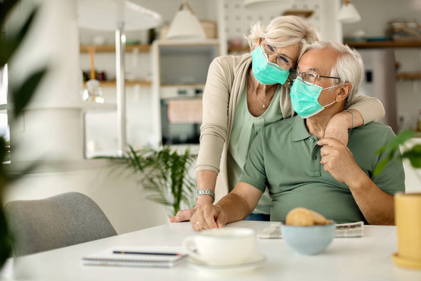 Αγκαλιασμένο ώριμο ζευγάρι που φοράει προστατευτικές μάσκες προσώπου ενώ χαλαρώνει στο σπίτι κατά τη διάρκεια επιδημίας ιού.  - Φωτογραφία, εικόνα