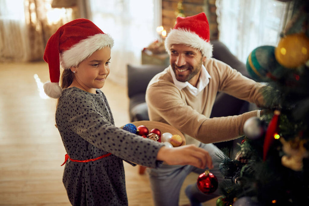 Ευτυχισμένο κοριτσάκι απολαμβάνει στη διακόσμηση χριστουγεννιάτικο δέντρο με τον πατέρα της στο σπίτι. - Φωτογραφία, εικόνα