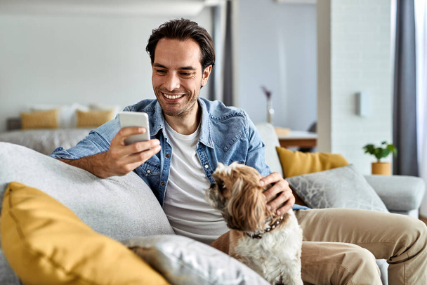 Young happy man text messaging on cell phone while sitting on the sofa with his dog. - Foto, Bild