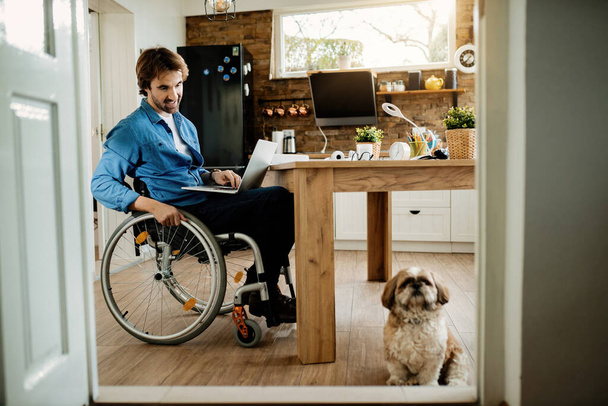 Szczęśliwy biznesmen na wózku inwalidzkim patrząc na swojego psa podczas pracy na laptopie w domu. - Zdjęcie, obraz