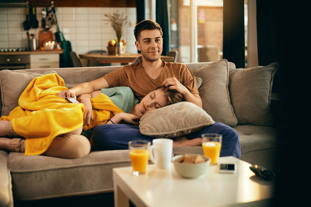 Young smiling man watching a movie while girlfriend is napping on his lap in the living room.  - Photo, Image