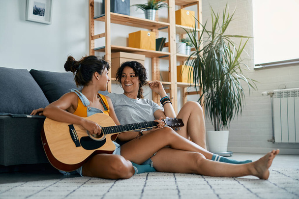 Молоді щасливі жінки розмовляють під час гри на акустичній гітарі і відпочивають на підлозі вдома
. - Фото, зображення