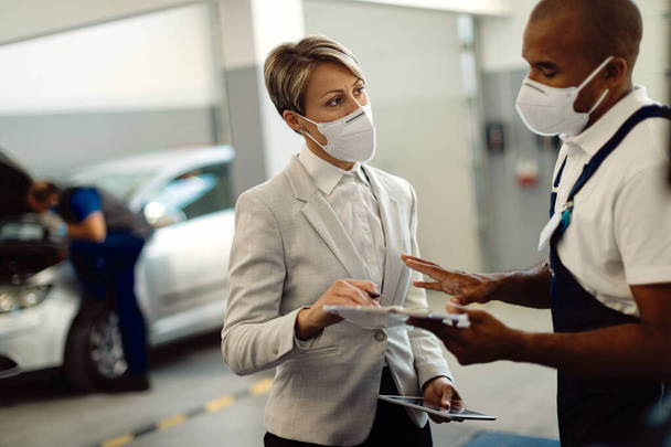 Businesswoman and African American repairman wearing protective face masks while talking and going through reports in car service workshop.  - Foto, Imagem