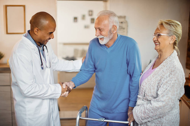 Happy African American doctor handshaking with senior man who is using a walker while being with his wife at nursing home.  - Photo, Image