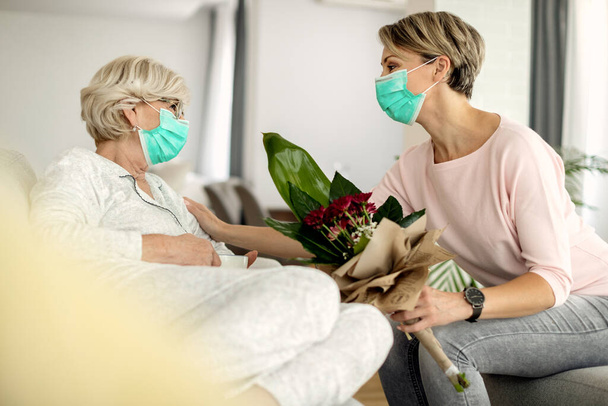 Happy woman talking to her mother while visiting her at home during coronavirus epidemic.  Both of them are wearing protective face masks.  - Photo, image