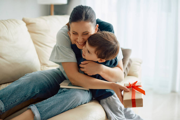 Маленький хлопчик дарує мамі подарункову коробку і обіймає її вдома
.  - Фото, зображення
