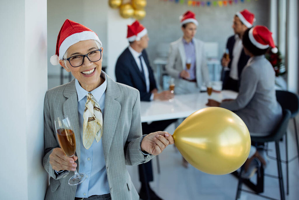 Щаслива зріла бізнес-леді, що тримає повітряну кулю і має склянку шампанського на Різдвяній вечірці в офісі. Її колеги на задньому плані..  - Фото, зображення
