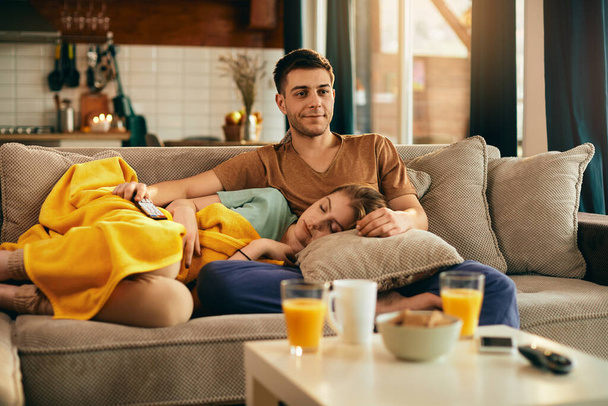 Young couple relaxing in the living room. Smiling man is watching TV while woman is sleeping n the sofa.  - Fotografie, Obrázek