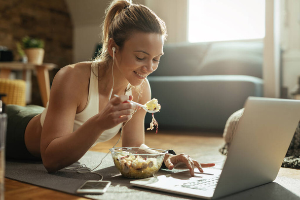 Young happy sportswoman eating salad and using laptop while relaxing on the floor at home.  - Foto, imagen