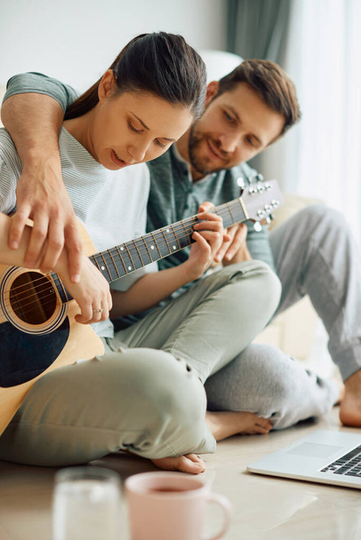 Young man teaching his wife to play acoustic guitar at home. Focus is on woman.  - 写真・画像