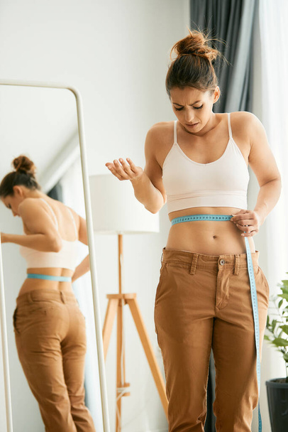 Young woman feeling dissatisfied with her weight loss while measuring her waist with tape measure at home. - Zdjęcie, obraz