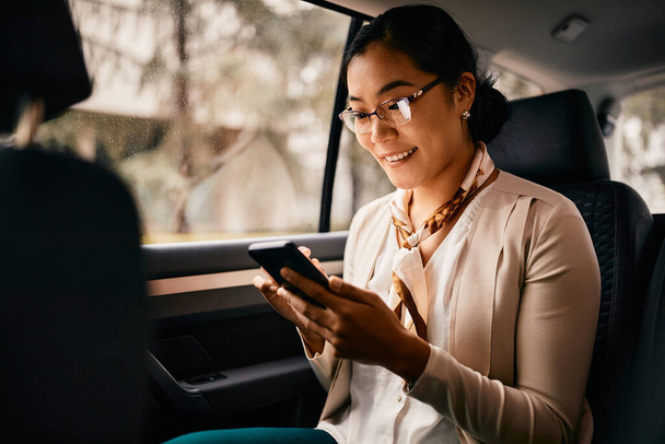 Smiling Asian female CEO sitting on the backseat and using smart phone while going on a business trip by car. - Foto, Imagen