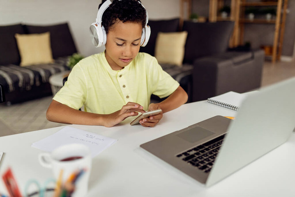 Smiling African American kid wearing headphones and text messaging while studying at home.  - Photo, image