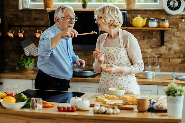 Happy mature man giving his wife to try out food he is cooking in the kitchen.  - 写真・画像