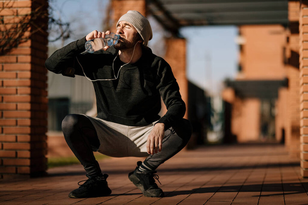 Young athletic man crouching on the street while drinking fresh water from a bottle.  - 写真・画像