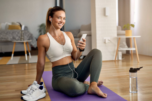 Happy athletic woman text messaging on smart phone while resting on the floor in the living room.  - Fotografie, Obrázek