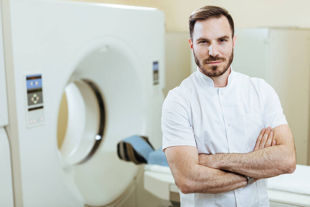 Pewny siebie radiolog ze skrzyżowanymi ramionami stojący przy skanerze MRI w klinice i patrzący w kamerę.  - Zdjęcie, obraz