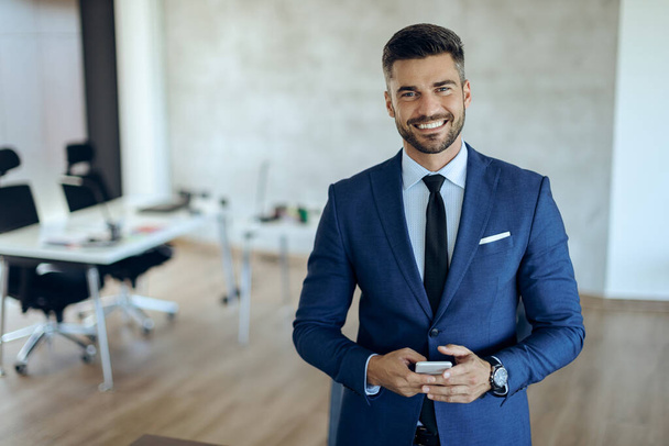 Portrait of young happy entrepreneur holding mobile phone and looking at camera while standing in the office.  - Fotoğraf, Görsel
