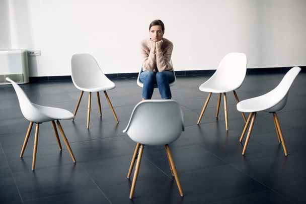 Displeased woman sitting in a chair and thinking while waiting for the group therapy to start.  - Foto, Imagen