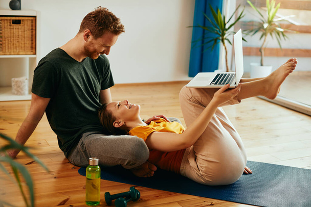 Happy couple talking and having fun while using laptop after sports training at home. - Фото, изображение