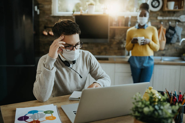 Distraught entrepreneur having problems while working on a computer at home and wearing protective face mask due to coronavirus pandemic.  - 写真・画像