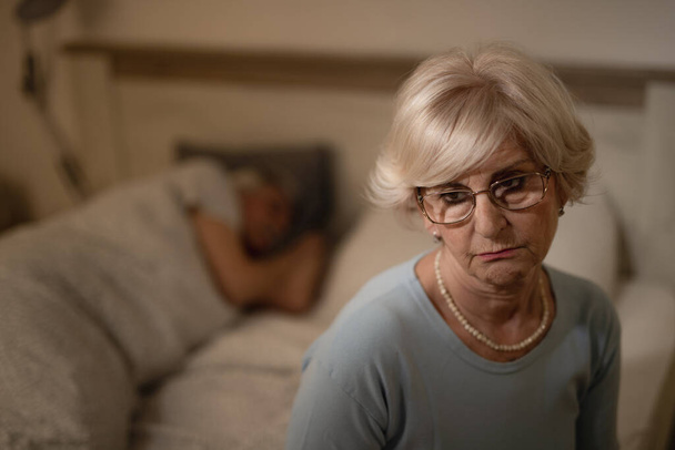 Penzitivní dospělá žena, která na něco myslí, zatímco v noci sedí v ložnici. Její manžel spí v pozadí.. - Fotografie, Obrázek