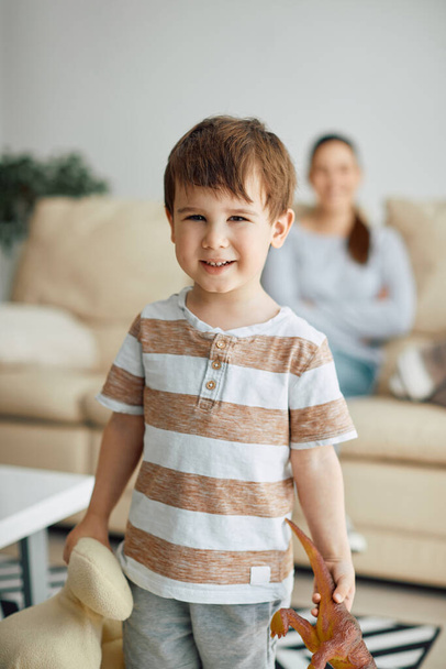 Happy little boy holding his toys and looking at camera at home. His mother is in the background.  - Foto, imagen