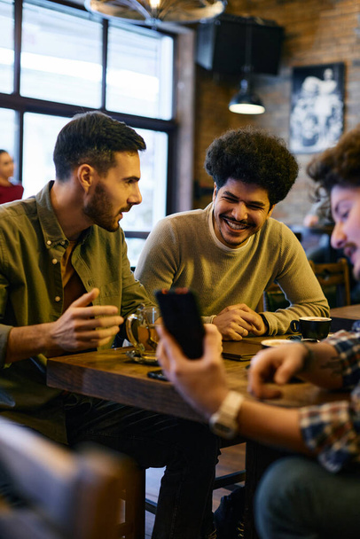 Group of happy male friends watching sports results on smart phone while sitting in a cafe, Focus is on Lebanese man. - 写真・画像