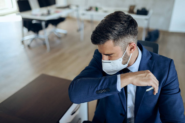 Male entrepreneur sneezing into elbow while wearing protective face mask in the office.  - Fotó, kép