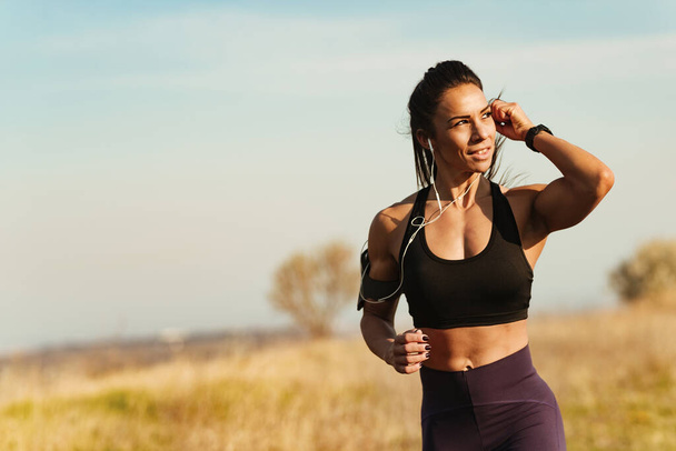 Młoda muskularna budowa kobieta dostosowuje słuchawki podczas biegania w naturze. Przestrzeń kopiowania.  - Zdjęcie, obraz
