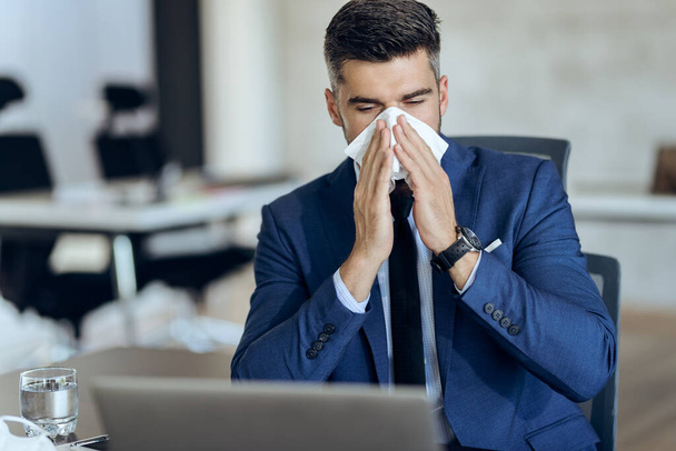 Businessman feeling sick and blowing nose while working at his office desk.  - Foto, imagen