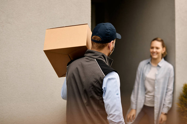 Back view of delivery man carrying cardboard box while delivering package to customer's home.  - Zdjęcie, obraz